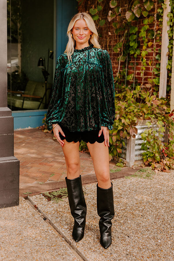 Woman wearing a luxurious green and black velvet burnout top with intricate patterns, styled with chic black scalloped shorts and knee-high boots, creating an elegant, evening-ready look.